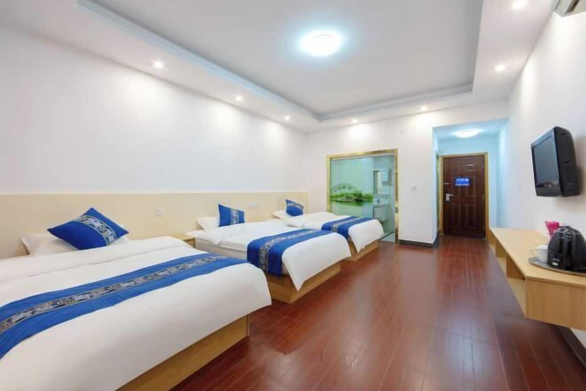 A hotel room with three beds, a TV, a desk, and a bathroom visible through a glass door.