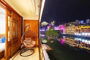 Una habitación con puerta de madera, una silla de mimbre y vistas de un río con edificios por la noche.
