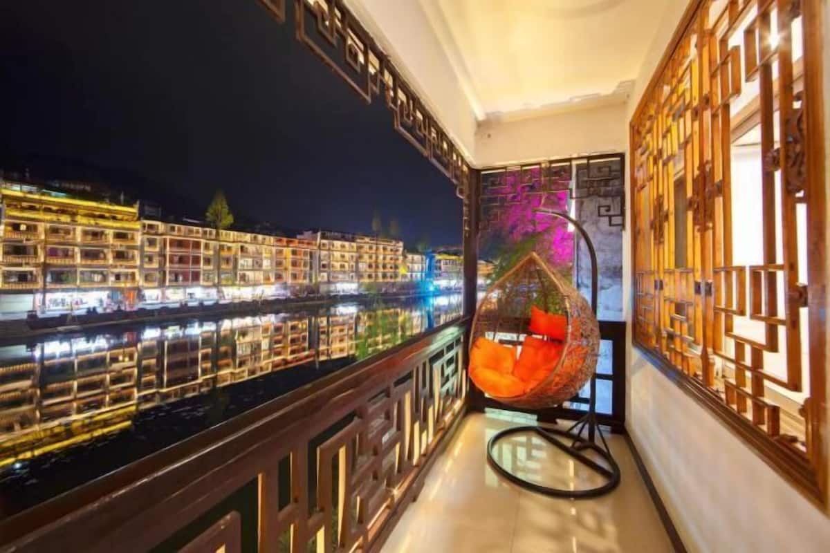 A balcony with a view of a river and buildings, a hanging chair, and traditional wooden lattice design.