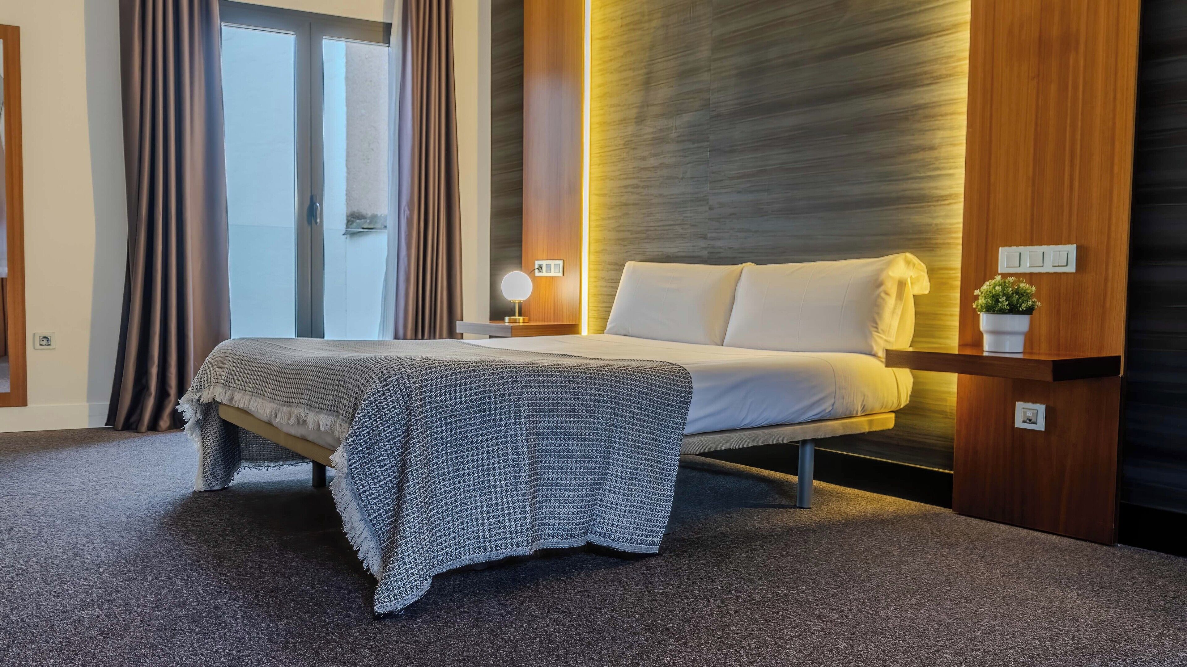 Una habitación de hotel moderna con una cama, una mesita de noche con lámpara, una planta pequeña y vistas al exterior.