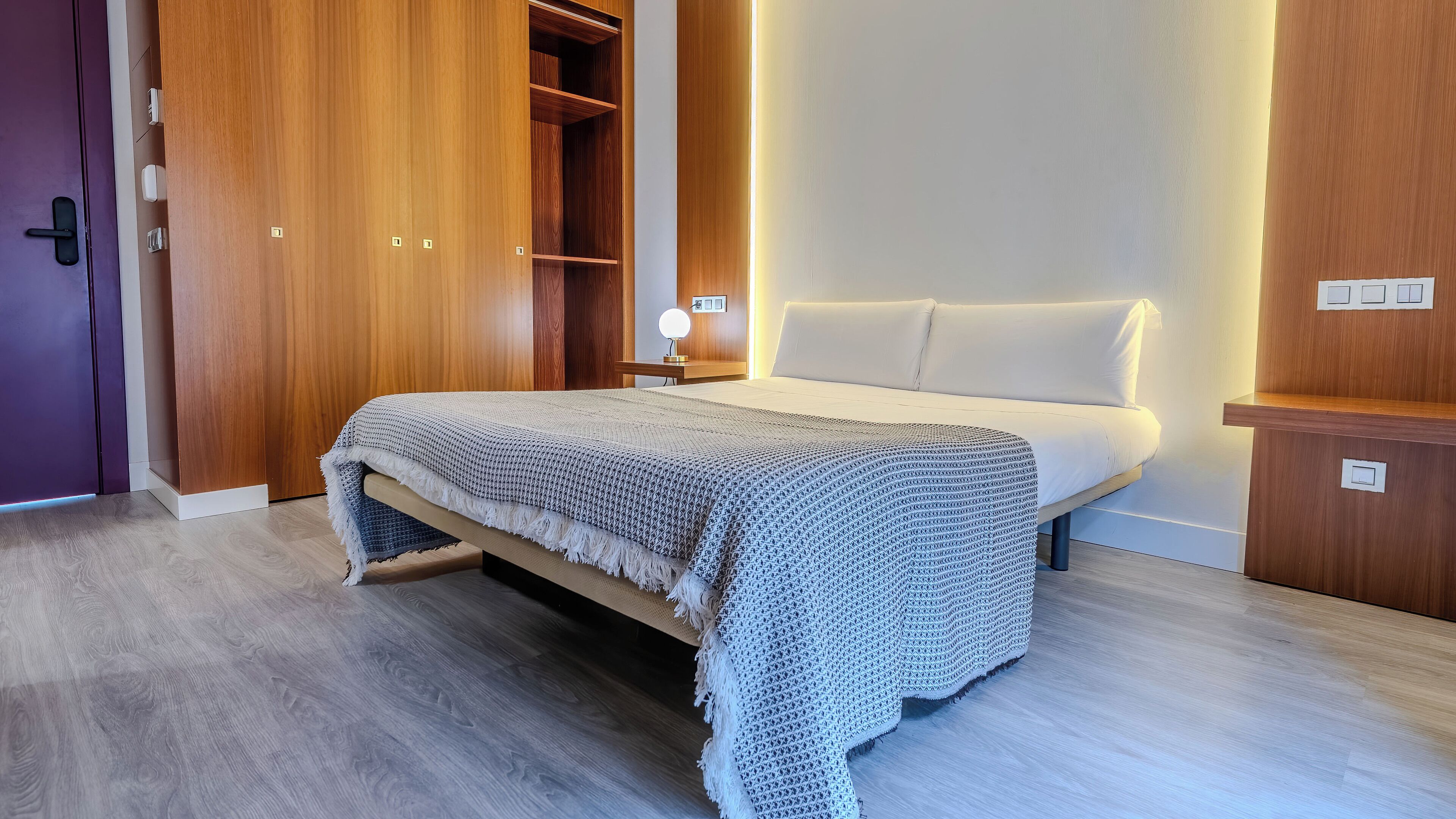 A modern bedroom with a bed, wooden wardrobe, and bedside table with a lamp.