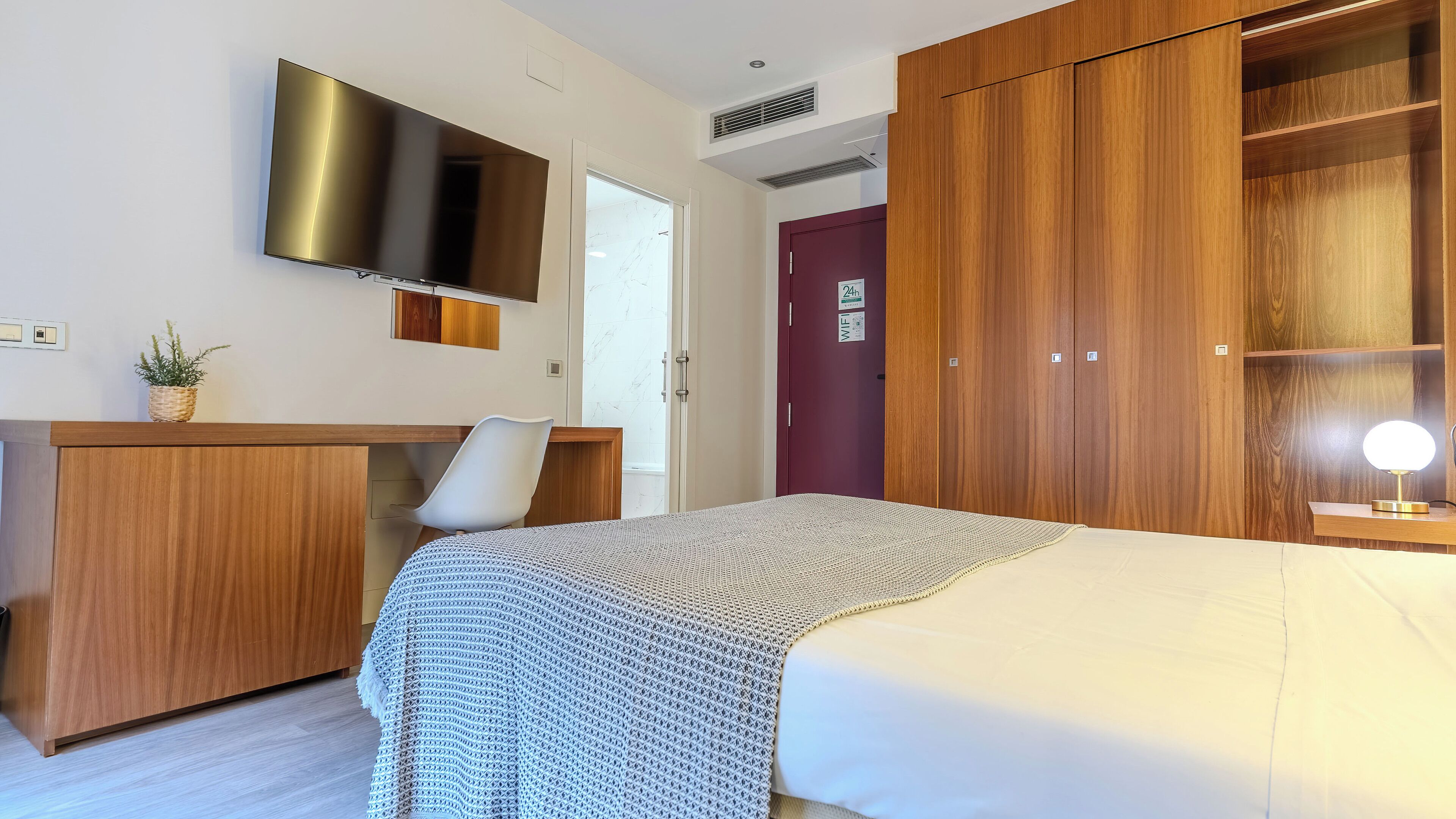 Una habitación de hotel con cama, escritorio, televisi ón, armario y lámpara de noche.
