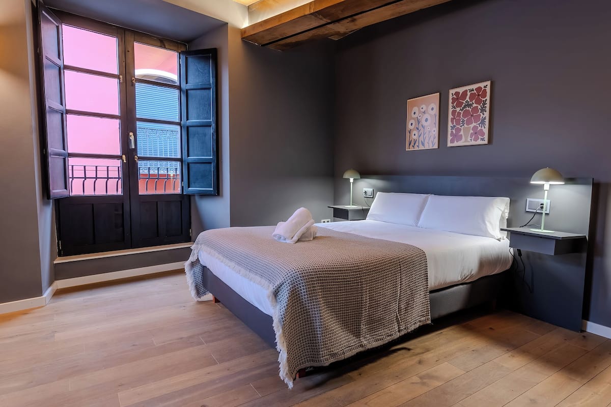 Un dormitorio con cama, mesitas de noche, una ventana con persianas y una obra de arte en la pared.