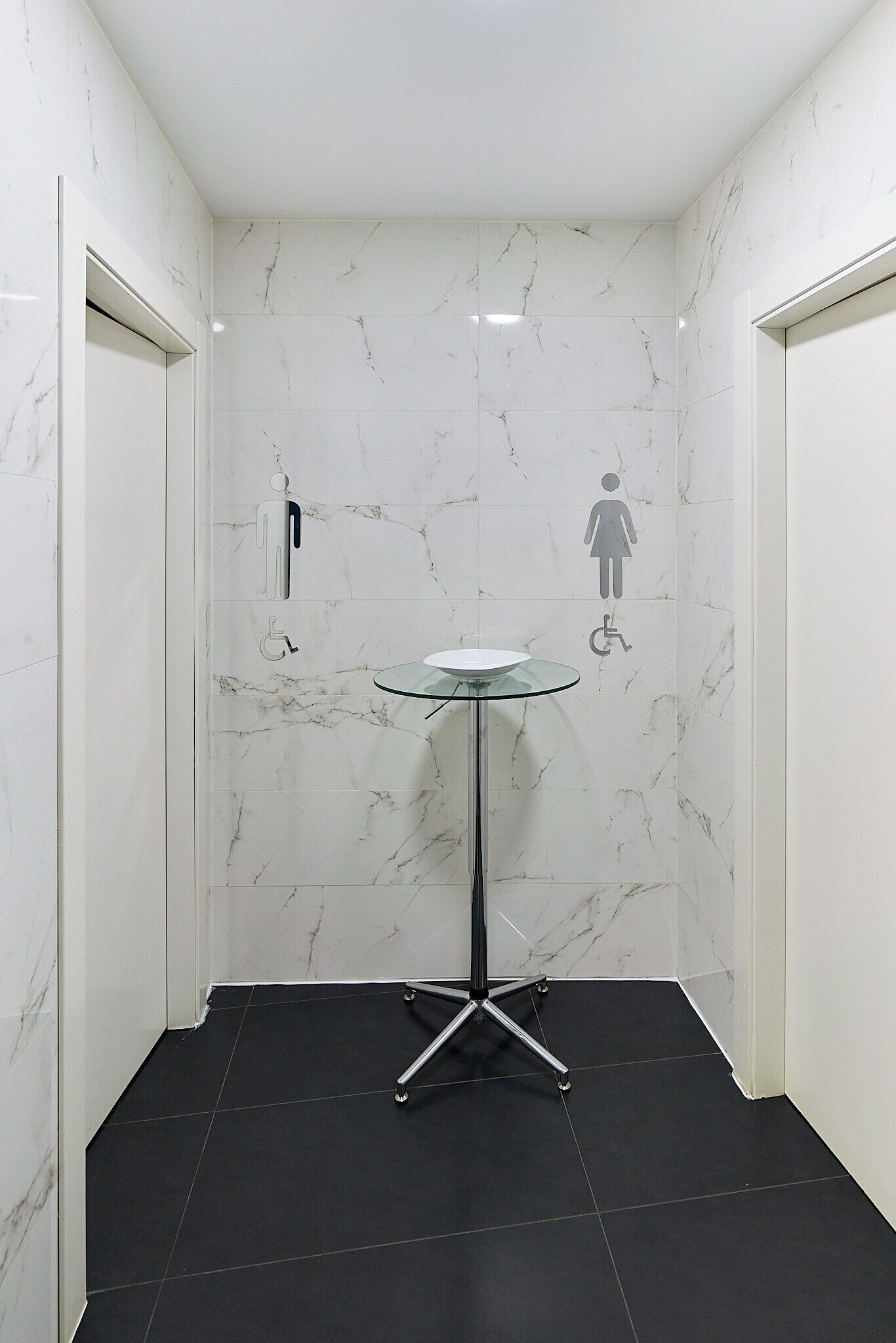 A small, modern bathroom with marble walls, a glass sink, and a black floor.