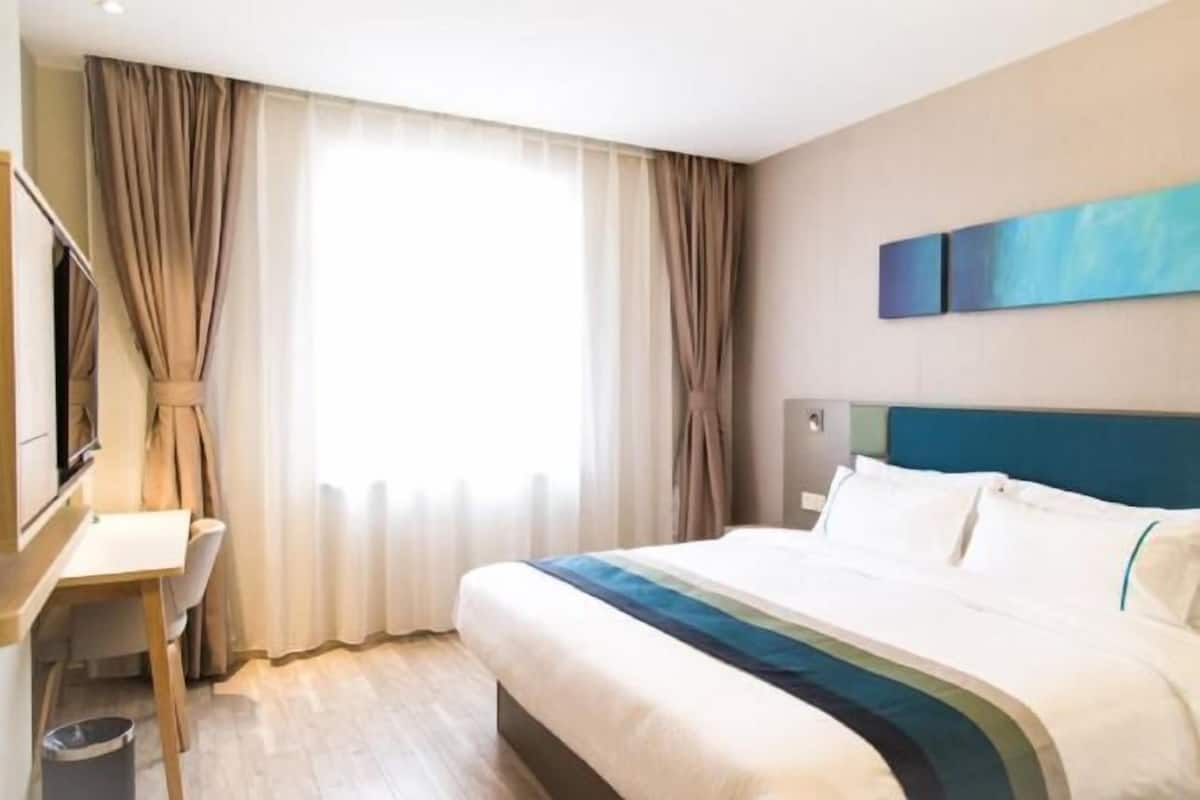 Una camera d'albergo con un letto, una scrivania, una sedia, un cestino e una finestra con tende.