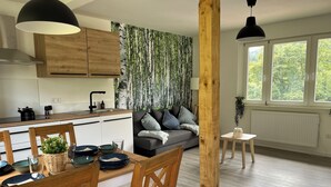 Una cocina moderna con armarios de madera, una mesa de comedor con sillas, y una sala de estar con sofá y vistas a árboles a través de amplios ventanales.
