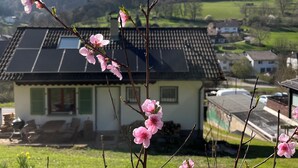 Una casa con techo inclinado y paneles solares, rodeada de un paisaje verde y un cielo despejado.