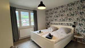 Un dormitorio con cama, ventana con cortinas, radiador, una mesita y pared con papel tapiz floral.