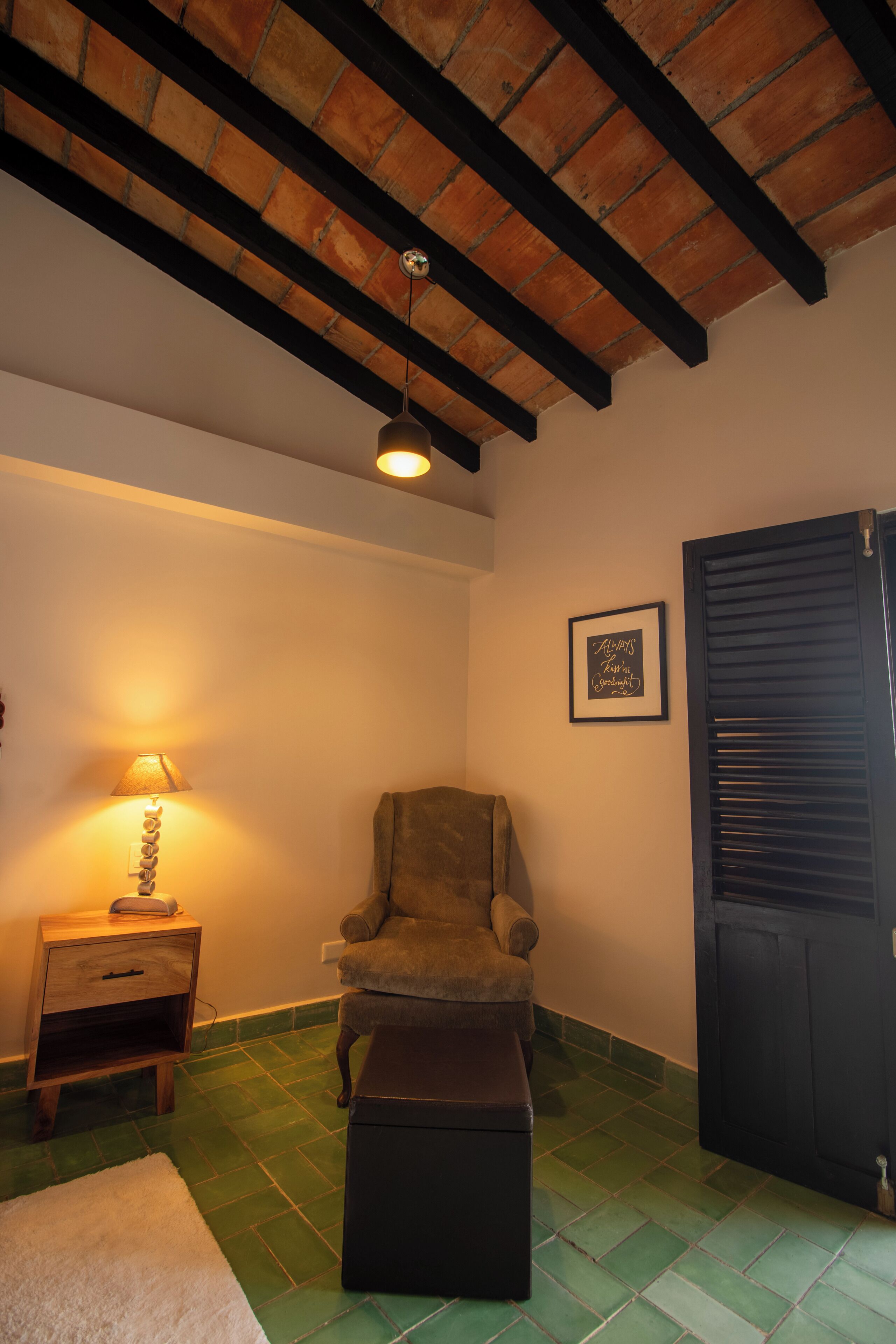 A room with a green tiled floor, a wooden bench, a chair, a bedside table with a lamp, a framed sign on the wall, and a dark door.