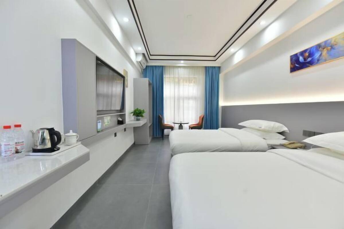 A hotel room with two beds, a TV, a small table with chairs, a plant, and a painting on the wall.
