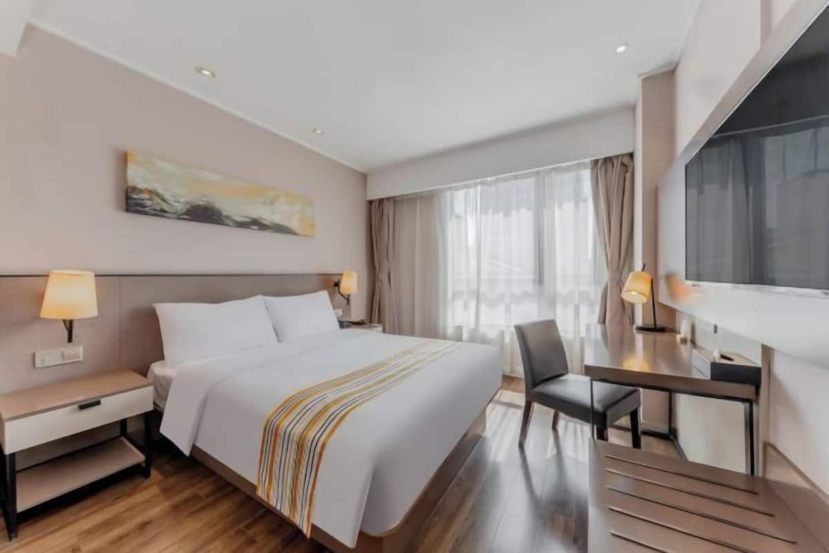 A hotel room with a bed, bedside table, lamp, desk, chair, and a large window with curtains.