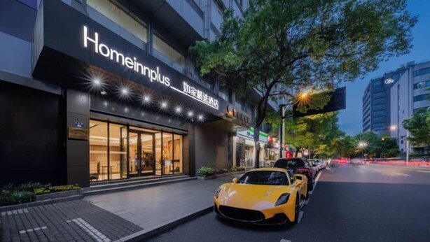 Un hotel llamado Homeinplus con una fachada moderna, un coche deportivo amarillo estacionado adelante y una calle bordeada de árboles.