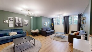 Un salon moderne avec un canapé bleu, un canapé gris, une table ronde et une table basse. Des tableaux encadrés ornent le mur, et une fenêtre est agrémentée de rideaux.
