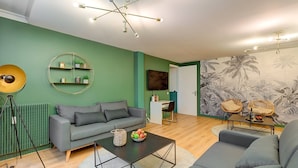 Un salon moderne avec un canapé gris, une table basse sur laquelle sont disposés des fruits, un miroir rond et un mur orné d’une fresque aux motifs tropicaux.