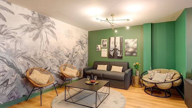 Un salon avec un mur d’accent vert, une grande fresque tropicale, un canapé gris, deux fauteuils en osier, une table basse et un fauteuil suspendu rond.
