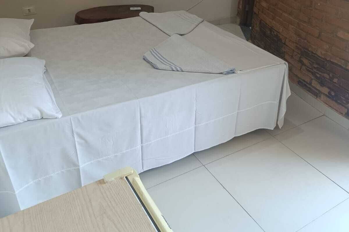 Ένα δωμάτιο με ένα μονό κρεβάτι, λευκό κλινοσκεπάσμα, ξύλινο προσκέφαλο και έναν τοίχο από τούβλα.