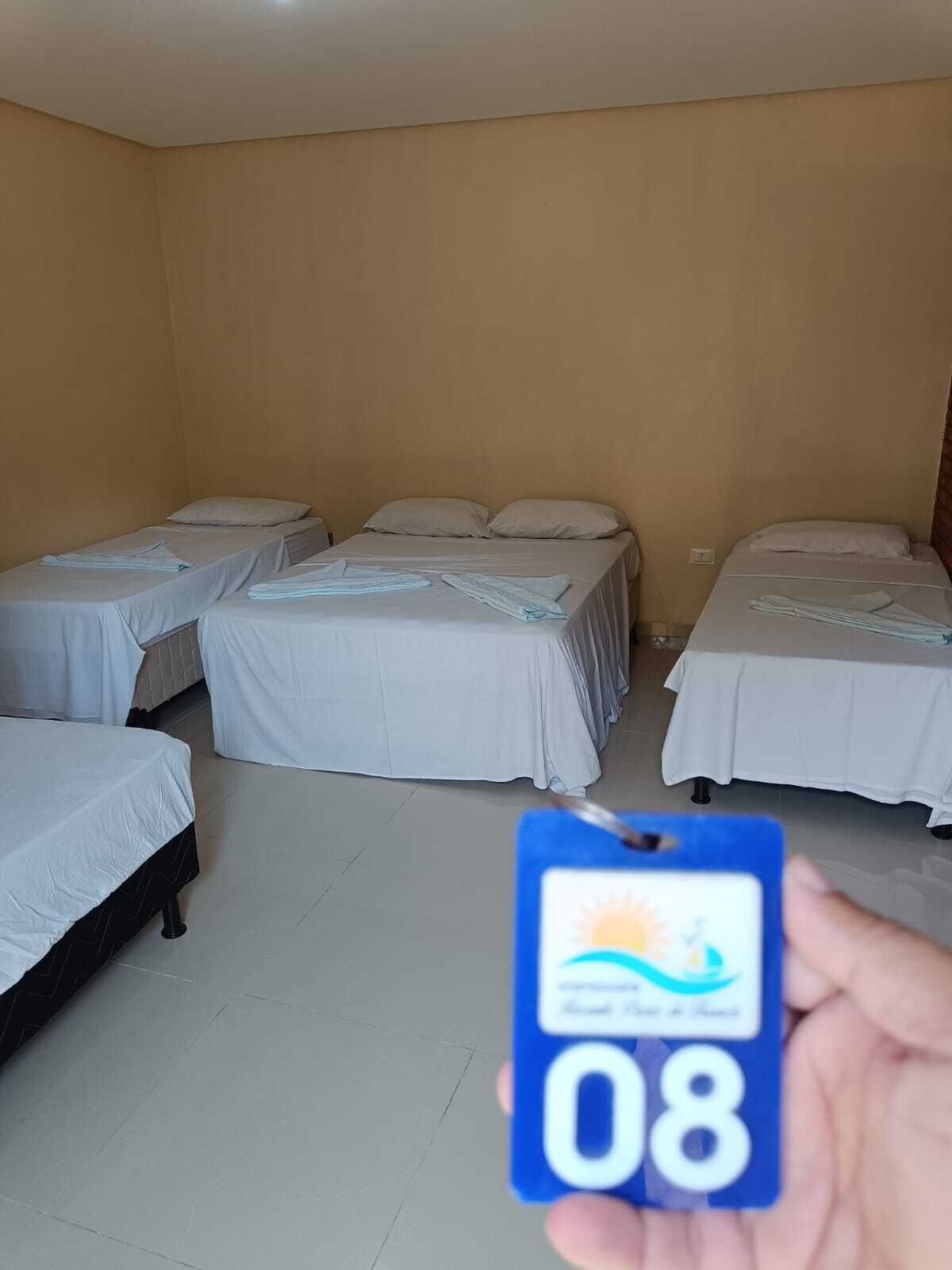 Une chambre d’hôtel avec quatre lits alignés, un sol carrelé et une main tenant une carte bleue portant le numéro 08.