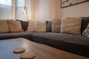 A modern living room with a sectional sofa, patterned throw pillows, a side table with coasters, and a floor lamp.