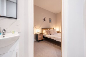 A hotel room with a white sink, a bedside table with a lamp, a bed with patterned pillows, and two framed pictures on the wall.