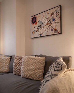 Ein modernes Wohnzimmer mit einem grauen Sofa, gemusterten Zierkissen und einem farbenfrohen, abstrakten Gemälde an der Wand.