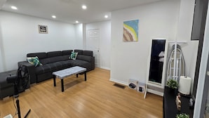 A modern living room with a sectional sofa, a coffee table, a standing fan, and a window with a view of the outside. - Clean precious house (Ajax)