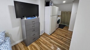 A small, well-lit room with a wooden floor, a grey cabinet with drawers, a flat-screen TV mounted on the wall, a white refrigerator, and a microwave on top of the fridge. - Clean precious house (Ajax)