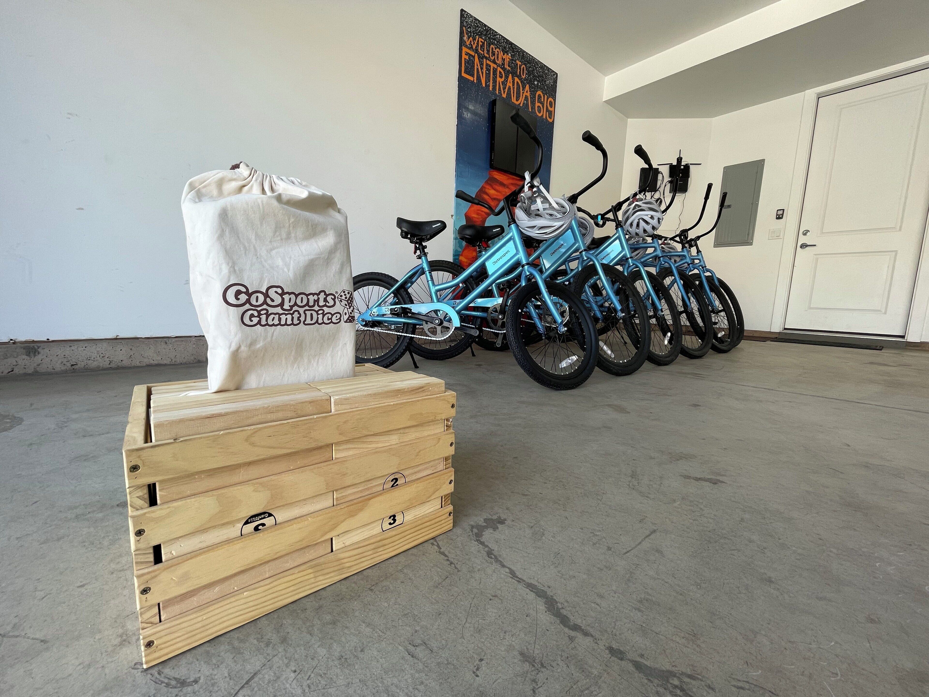 A row of bicycles parked indoors, a wooden crate with a bag labeled "CoSports Giant Dice," and a wall with a sign reading "Welcome to Entrada 619."