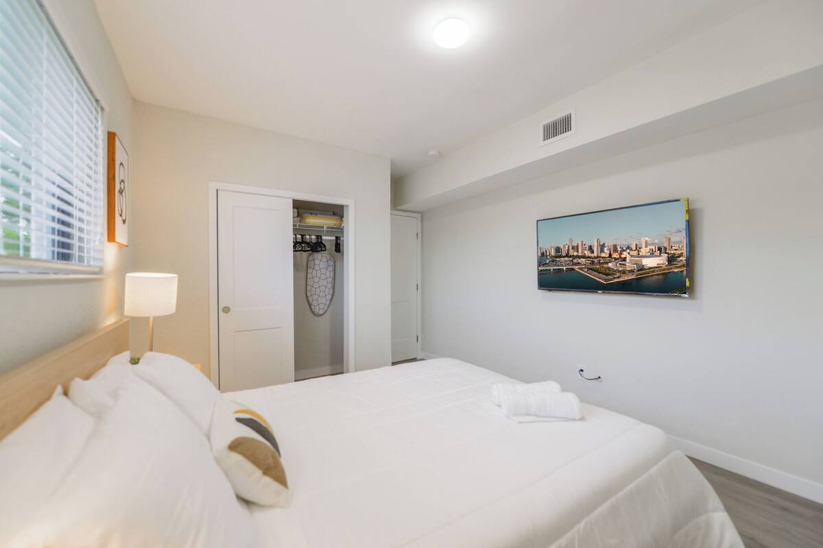 A hotel room with a bed, a closet, a painting of a cityscape, and a window with blinds.