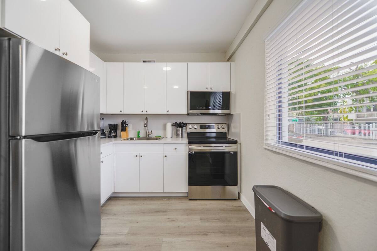 A compact kitchen with white cabinets, a stainless steel refrigerator, a microwave, an oven, and a window with blinds.