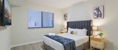 A modern hotel room with a large bed, a flat-screen TV mounted on the wall, a window with blinds, and a small bedside table with a lamp.