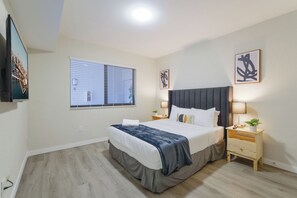 A modern hotel room with a large bed, a flat-screen TV mounted on the wall, a window with blinds, and a small bedside table with a lamp. - Stylish Apartments on Iconic Calle Ocho (Miami)