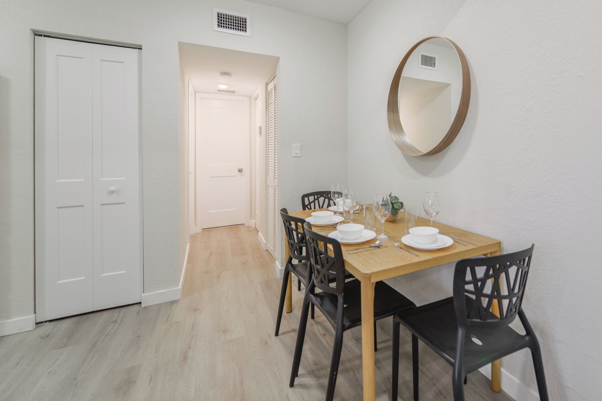 A dining area with a wooden table set for four, surrounded by black chairs. A round mirror hangs on the wall above the table.