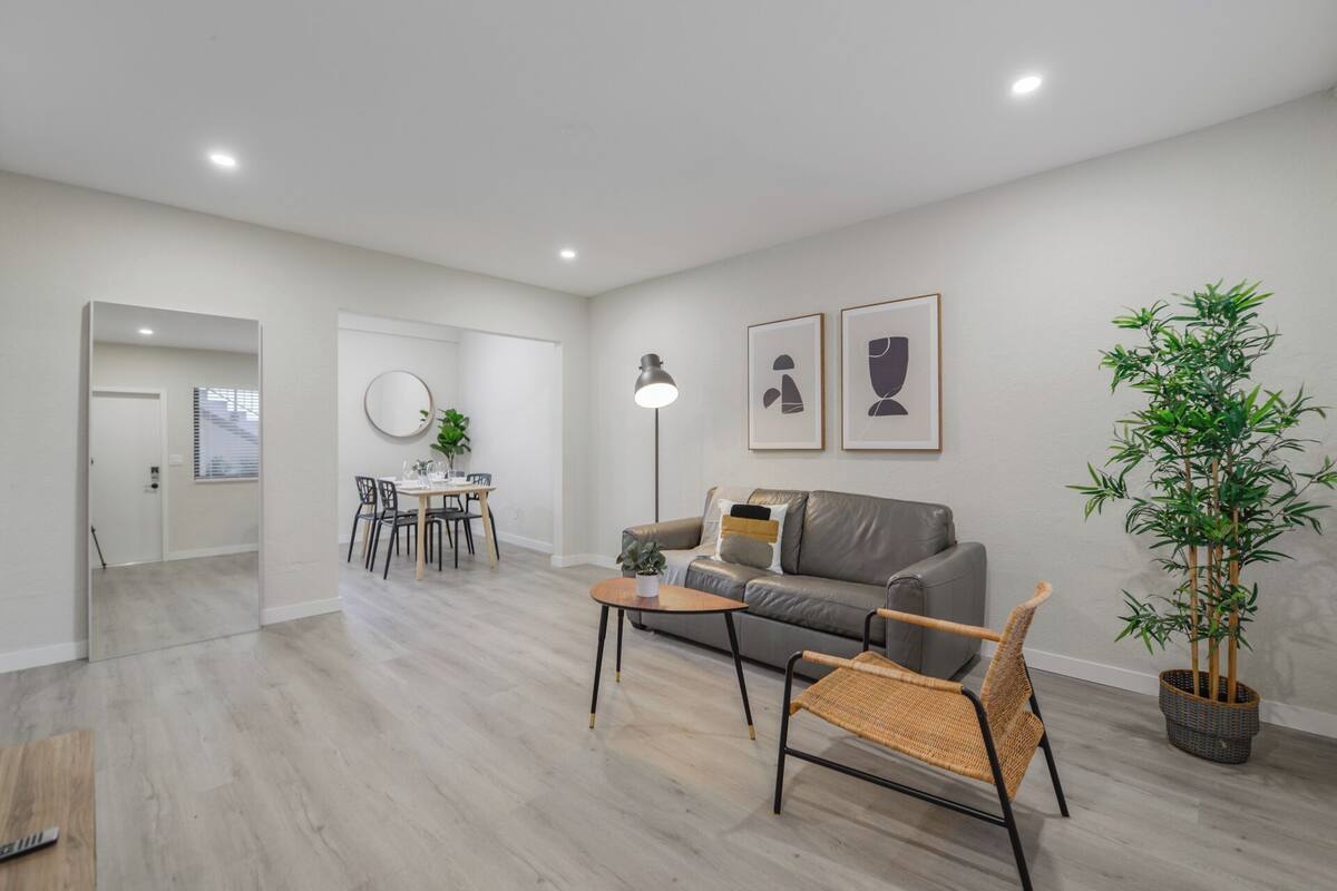 A modern living room with a grey sofa, a coffee table, and a dining area with a round table and chairs. There are two framed artworks on the wall and a potted plant.