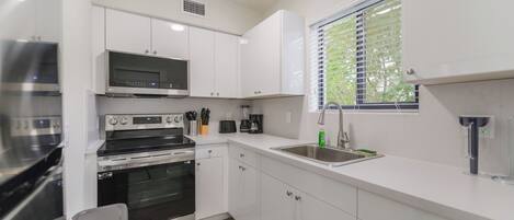 A compact kitchen with white cabinets, a microwave, a stove, a sink, and a window with blinds.