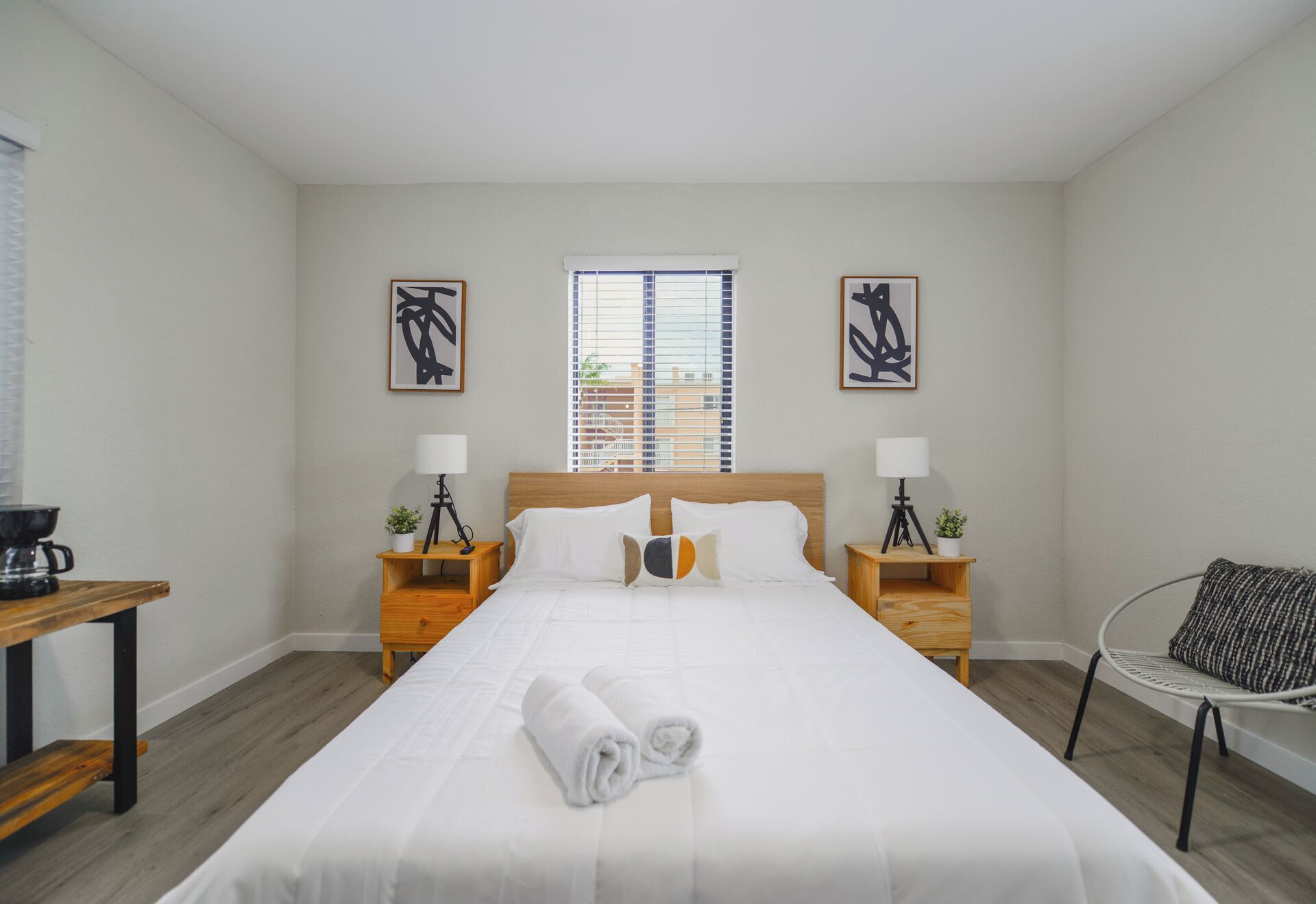 A neatly made bed with white linens and a rolled towel, two bedside tables with lamps and small potted plants, a window with blinds, and two abstract paintings on the walls.