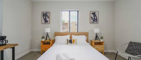 A neatly made bed with white linens and a rolled towel, two bedside tables with lamps and small potted plants, a window with blinds, and two abstract paintings on the walls.