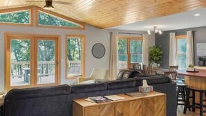 Una sala de estar acogedora con un sofá de color oscuro, una mesa de centro de madera y un comedor con una mesa y sillas de madera. Amplias ventanas con marcos de madera dejan entrar luz natural y ofrecen vistas al verde exterior.