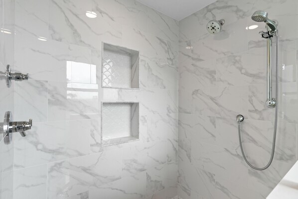 A modern bathroom with marble walls, a shower with a handheld showerhead, and a glass-enclosed window. - Beach Colony West 1C (Pensacola)
