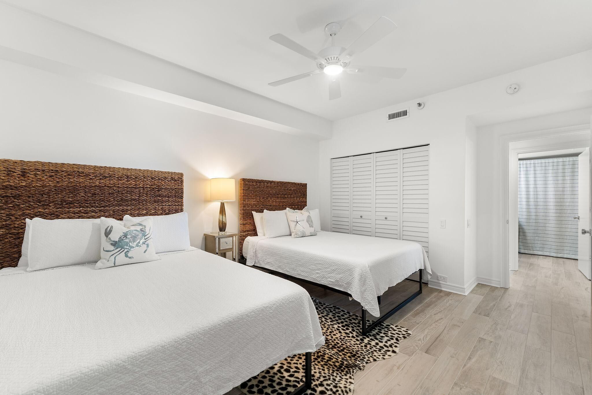 A hotel room with two beds, a ceiling fan, a nightstand with a lamp, a wardrobe, and a door leading to another room.