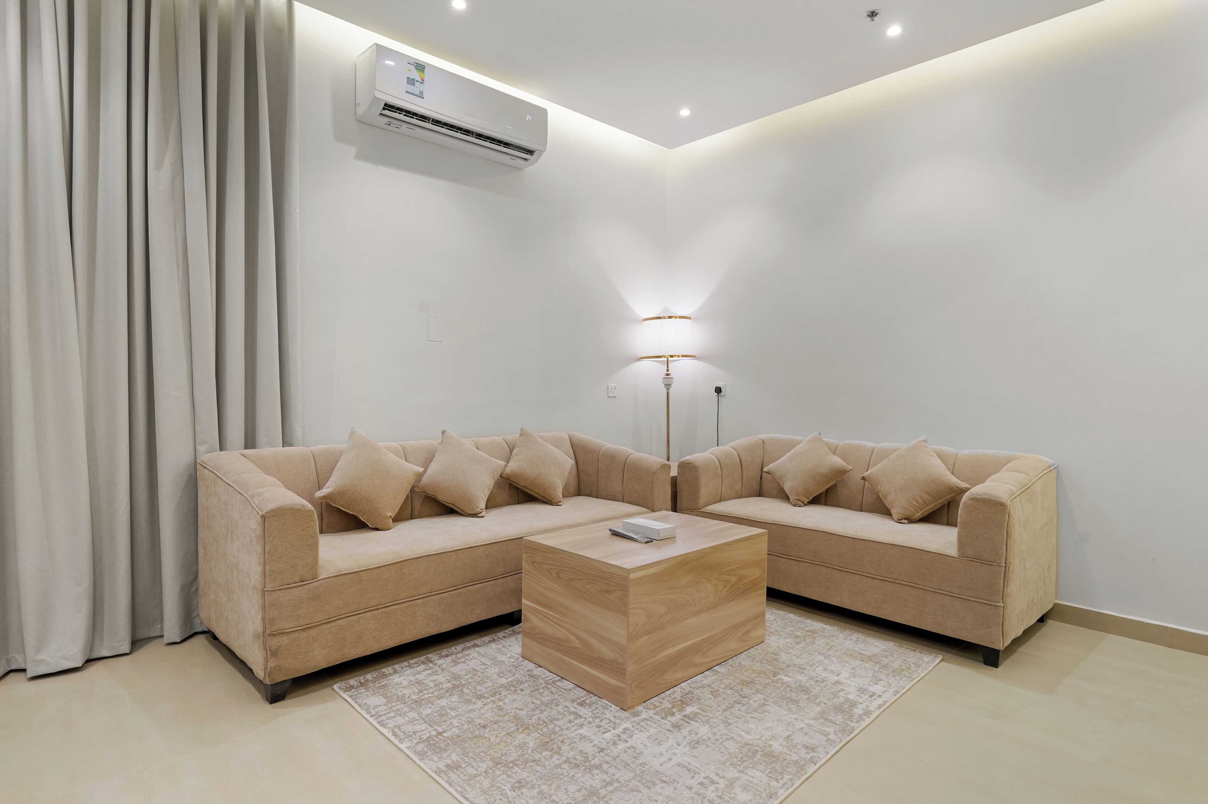 A modern living room with a beige sectional sofa, a wooden coffee table, a floor lamp, and an air conditioning unit on the wall.