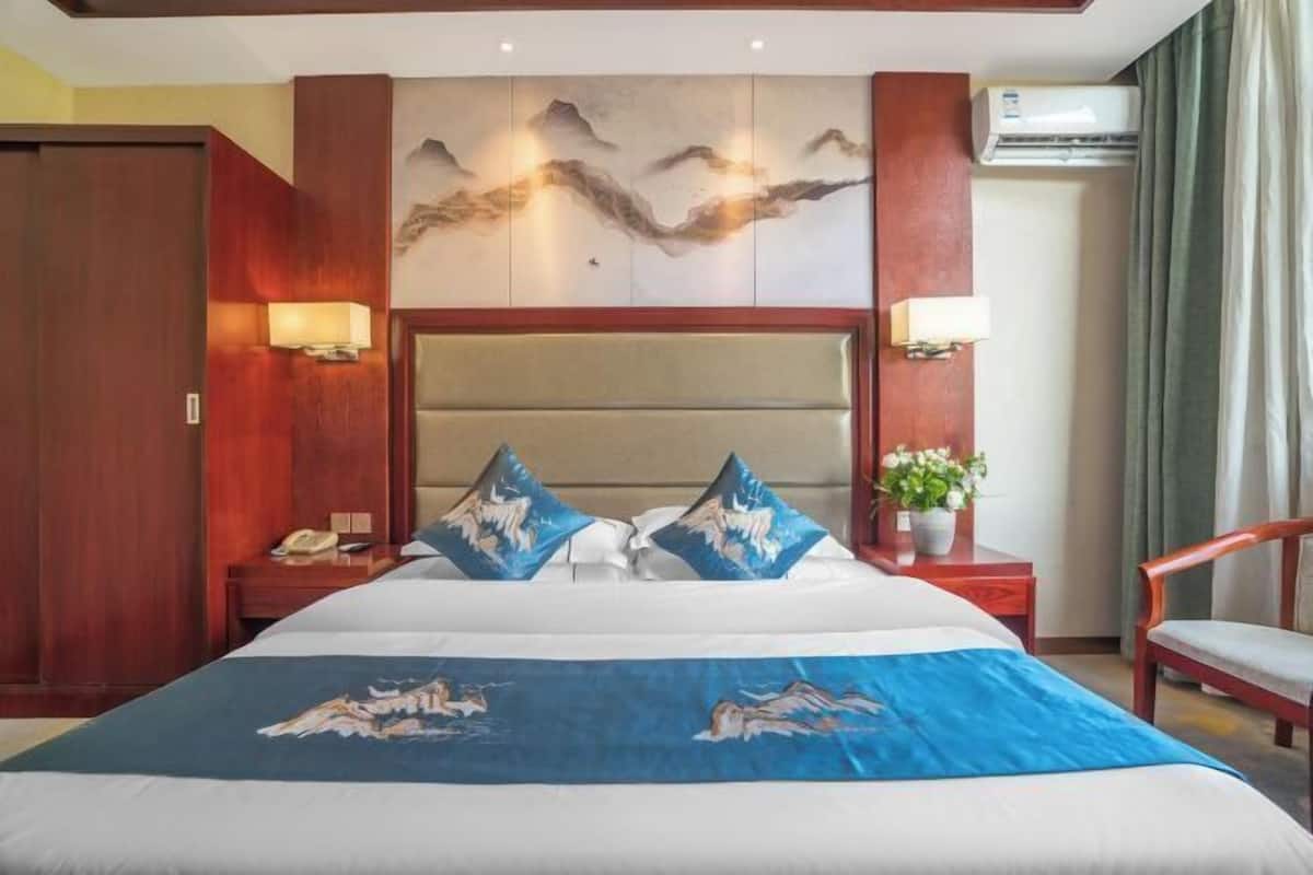 A hotel room with a large bed, wooden headboard, two bedside tables with lamps, a wooden chair, and a wall-mounted air conditioning unit.