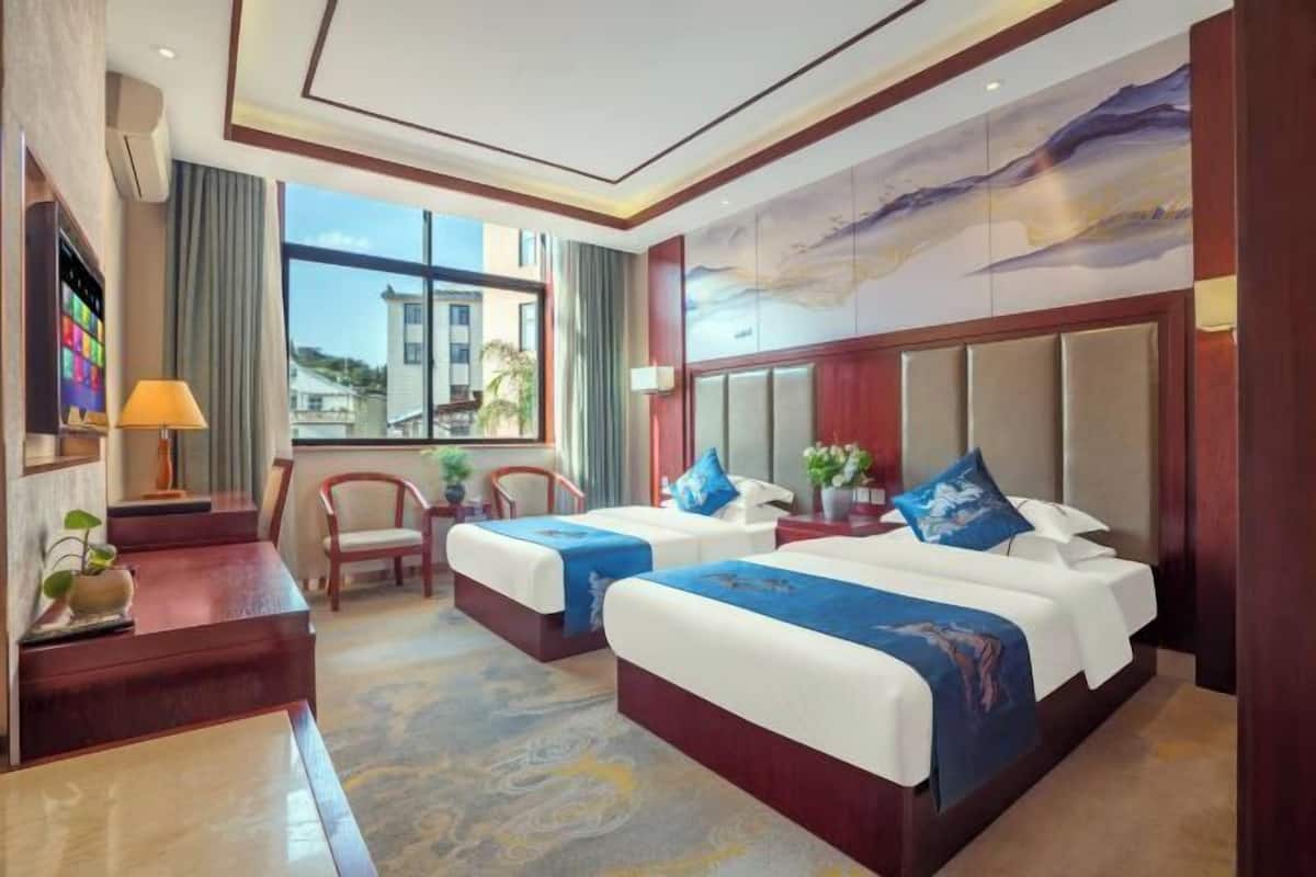 A hotel room with two beds, a desk, a chair, a TV, and a large window.