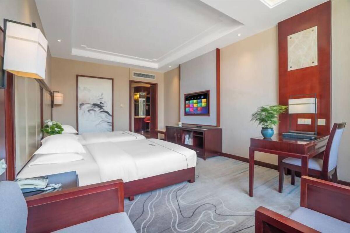 A hotel room with a large bed, a desk with a chair, a TV, a bookshelf, and a potted plant.