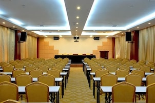 Un salón de conferencias con filas de sillas orientadas hacia un escenario con un atril, equipado con un proyector y una pantalla.