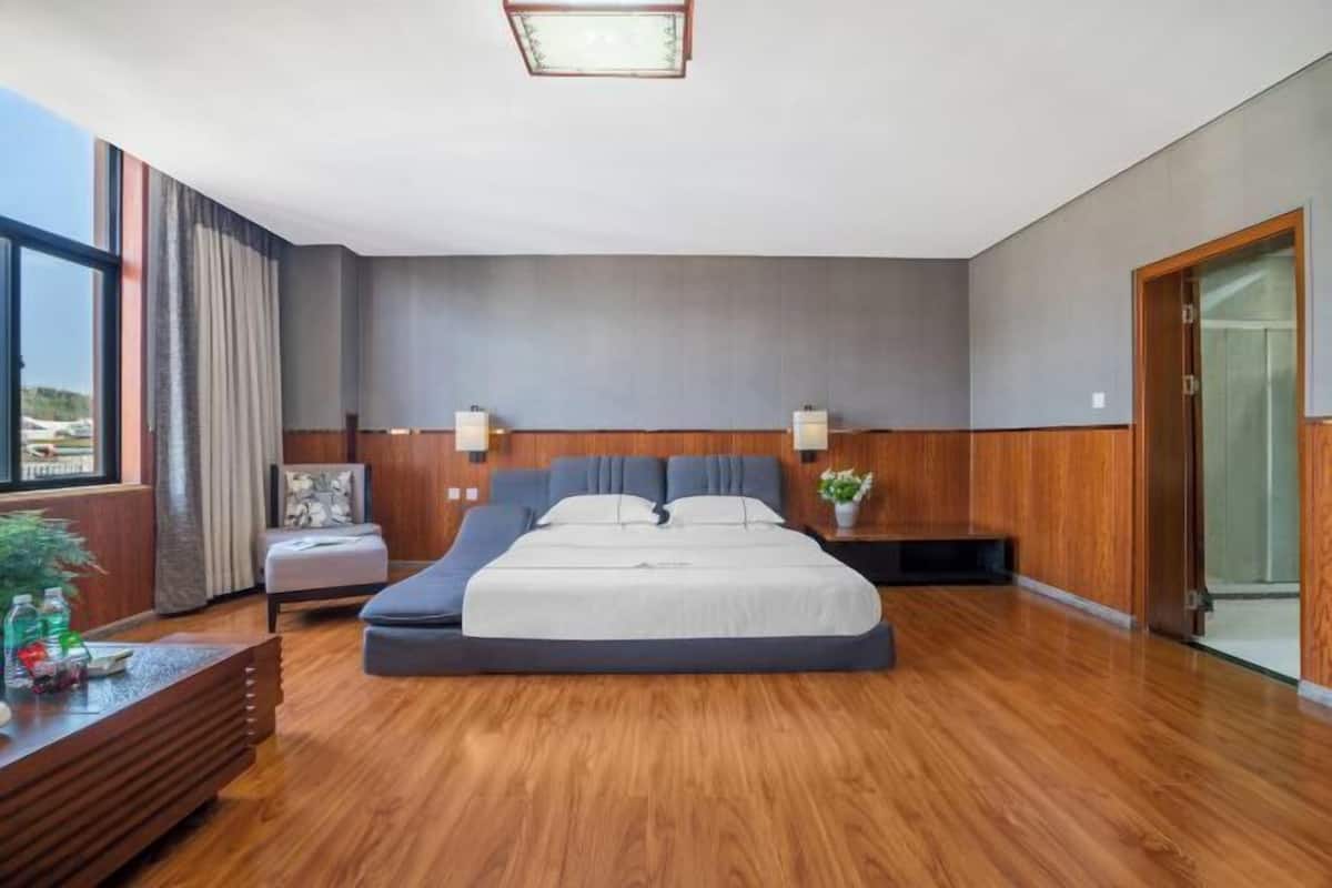 A modern bedroom with a large bed, wooden flooring, a nightstand, a chair, and a view of the outdoors.