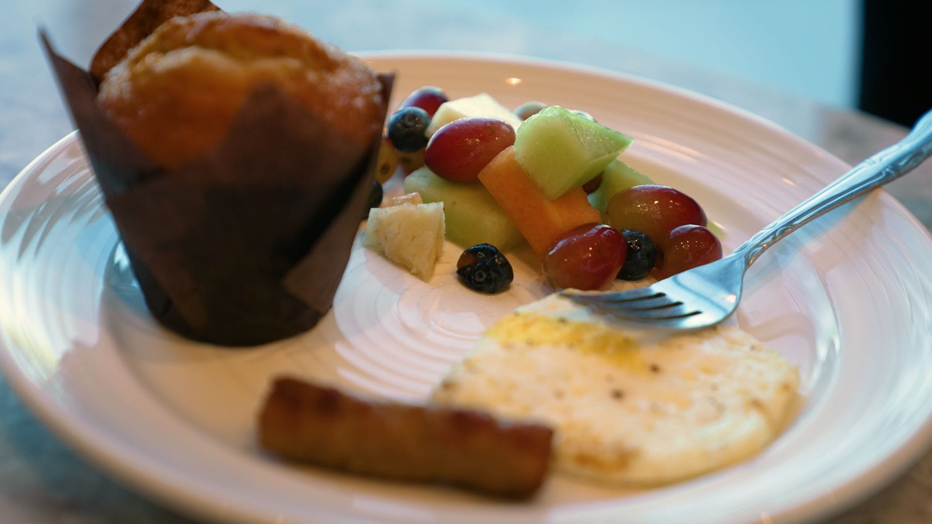 Een bord met een muffin, een sneetje toast en een fruitsalade met druiven, meloen en bosbessen.