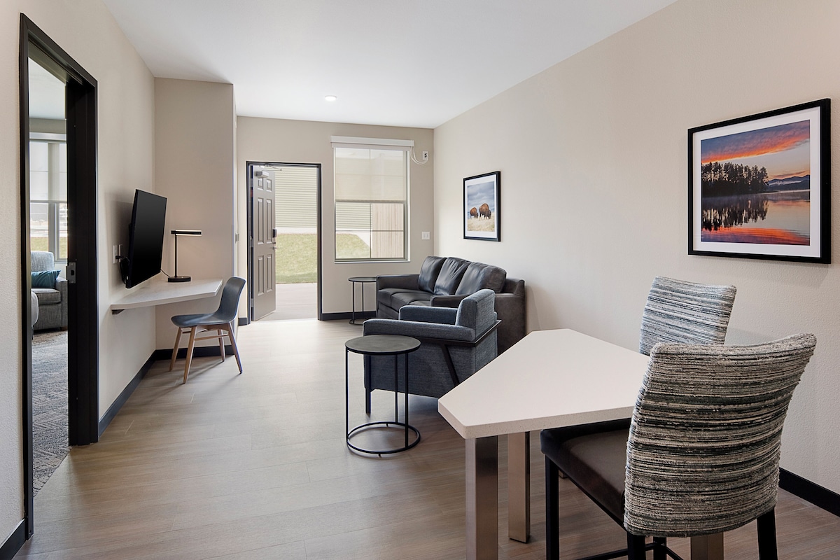 Un salon moderne avec un canapé, une table à manger et des chaises. Un téléviseur est fixé au mur, il y a une fenêtre avec des stores et un tableau encadré sur le mur.