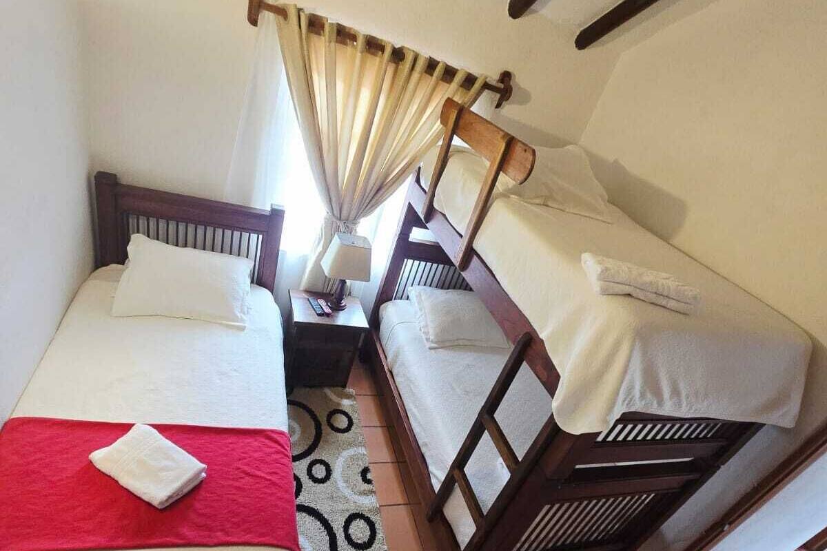 Una habitación pequeña y acogedora con dos camas de madera, una toalla roja, un tapete estampado y una ventana con cortinas.