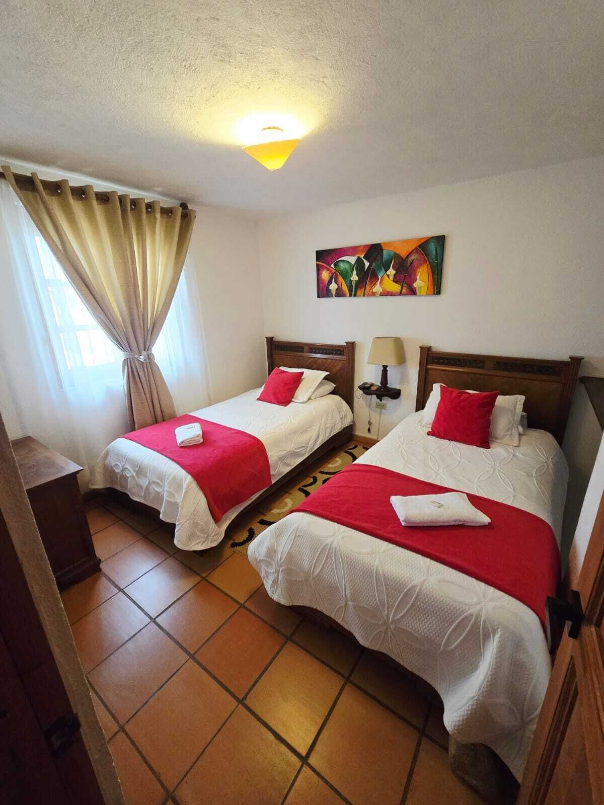 Une chambre d’hôtel avec deux lits, chacun agrémenté de draps rouges et blancs, un tableau au mur et une fenêtre avec des rideaux.