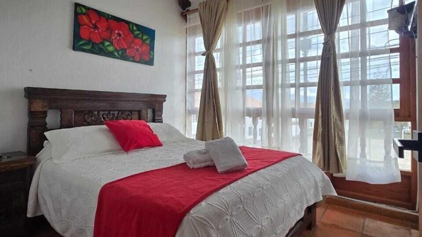 Une chambre à coucher avec une tête de lit en bois, un lit avec une literie blanche et rouge, un tableau représentant des fleurs accroché au mur et une fenêtre avec des rideaux.